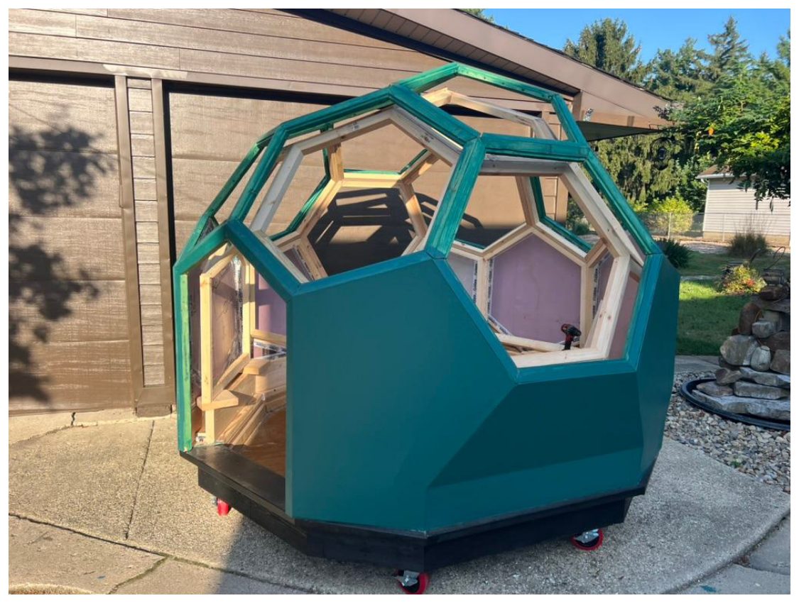 A geodesic dome shelter built from painted plywood panels with hexagonal windows, on casters in a driveway.